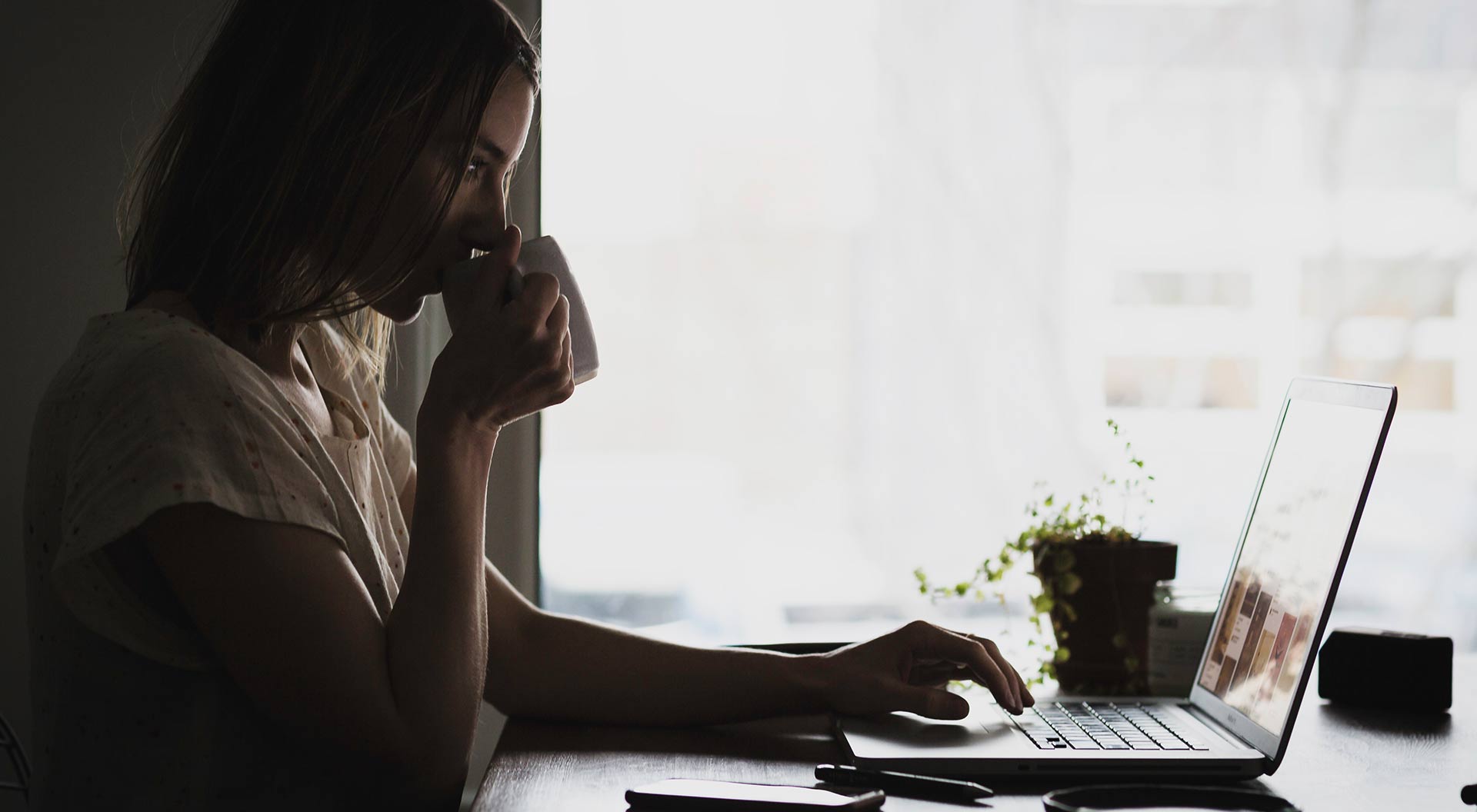 Woman working from home on laptop sipping coffee by a window