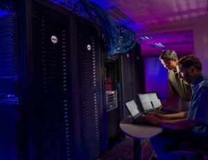 Two men using laptops in front of several racks filled with data servers and networking equipment